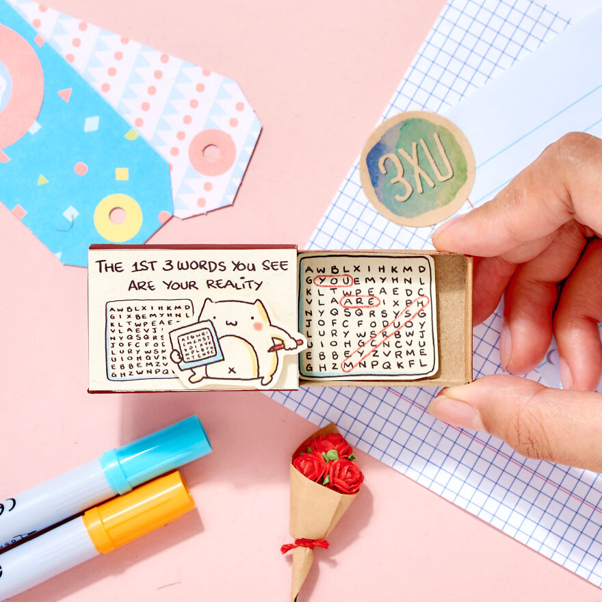 Hand holding a small puzzle box with a love word search, surrounded by craft supplies and a miniature bouquet of red roses.