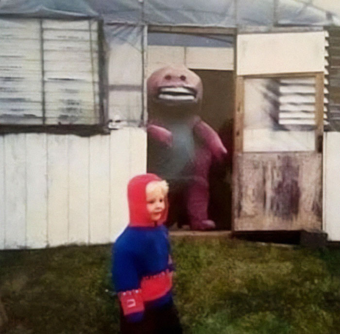 Child in a colorful hoodie walks outside, with a knockoff Barney character in a doorway behind them.
