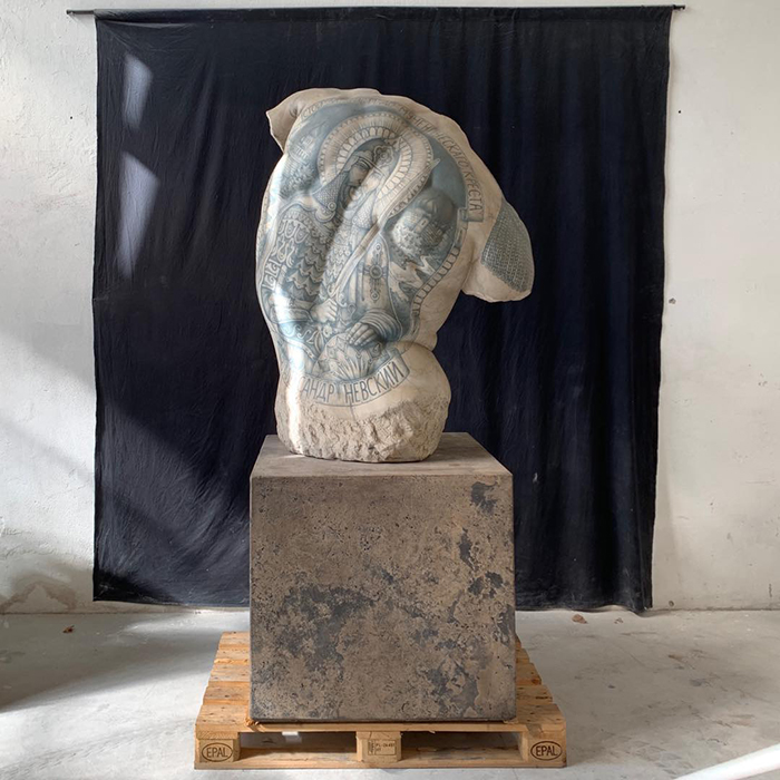 Sculpture with intricate tattoos by Italian artist displayed on a pedestal in front of a dark backdrop.