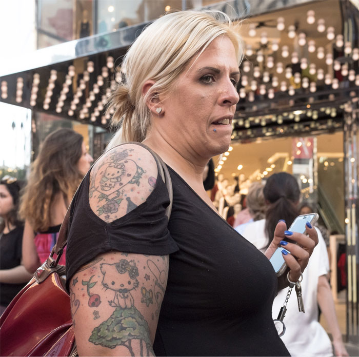 Woman with tattoos standing in a busy street, holding a phone and keys, showcasing her inked arm.