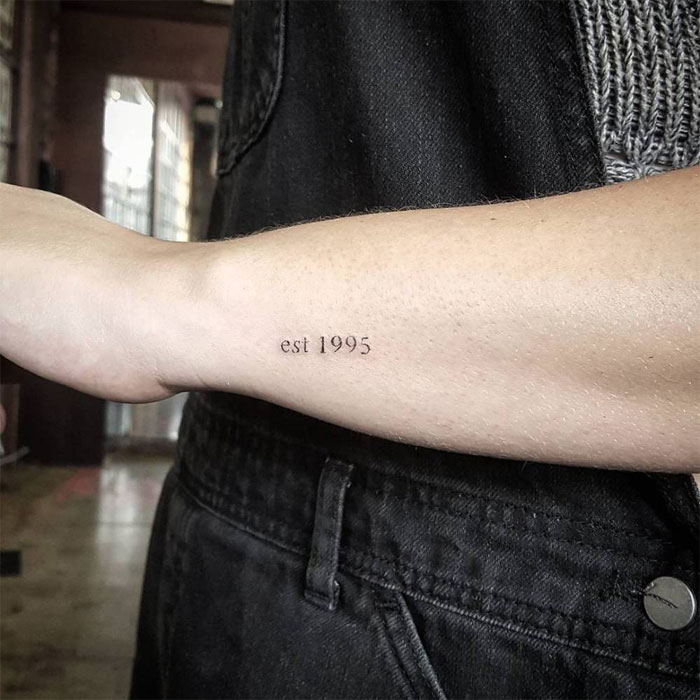 Forearm tattoo reading "est 1995" on a person in a black outfit, shared in a tattoo online group.