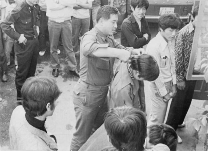 A South Korean Soldier Forcibly Cuts A Young Man's Hair In Front Of Others During A Nationwide Crackdown On Men With Long Hair And Women Wearing Short Skirts In South Korea - 1970s