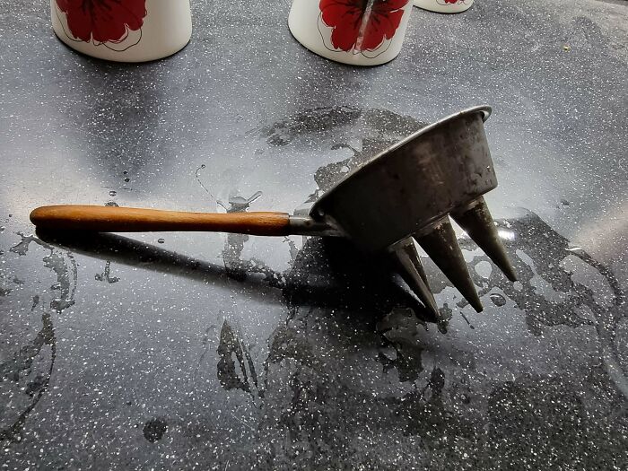 A Kitchen Utensil With A Wooden Handle, Attached To A Round Metal Cup With 3 Spikes (With Holes In)