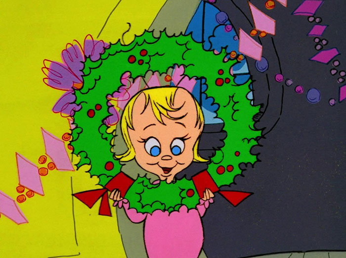 Cute animated character smiling through a colorful wreath with ribbons and flowers.