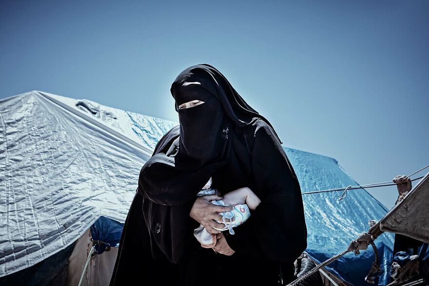 Woman in black niqab holding a baby outdoors near tents, captured by photographers showcasing beautiful women around the world