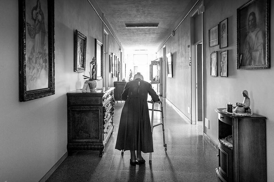 Black and white photo capturing an elderly woman walking with a walker in a long hallway, showcasing emotional portrait photography.