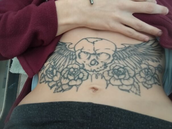 Skull tattoo with wings and roses on stomach, wearing maroon sweater, showcasing impressive new body art.