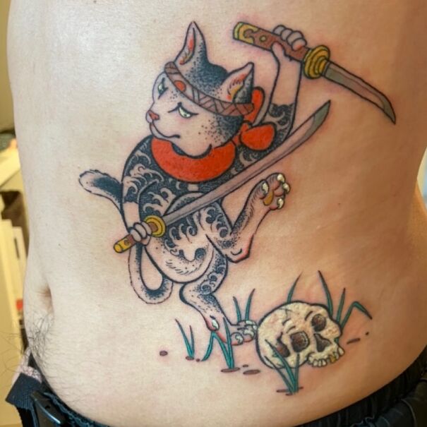Ninja cat tattoo with swords and a skull, shared by readers, showcasing impressive new body art.