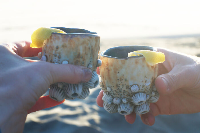 Hands holding nature-inspired ceramic cups with lemon peels near a beach setting.