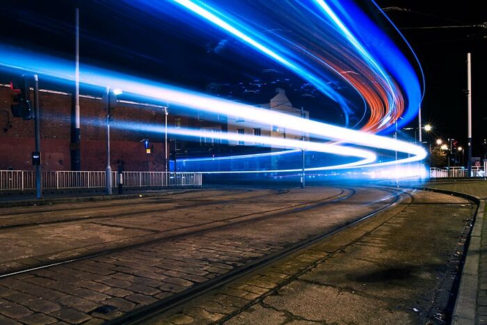 Long exposure of trams creating vibrant, otherworldly light trails on city streets at night.