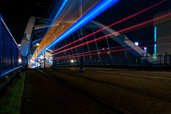 Long exposure photo of trams on a bridge at night, creating vibrant light trails.