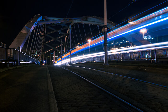 Long exposure photograph of a tram on a bridge at night, creating a surreal, otherworldly effect with streaking lights.