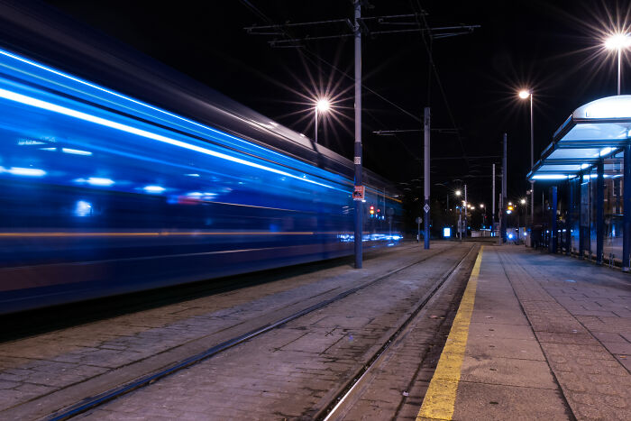 Long exposure of a tram at night, creating an otherworldly effect with blue light streaks on an empty city platform.