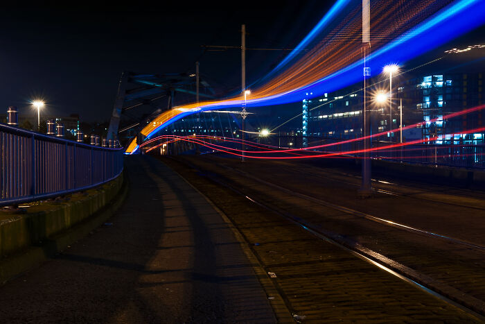 Long exposure photo of trams at night, creating streaks of light on a city bridge, resembling a scene from another world.
