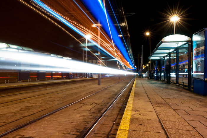 Long exposure shot of trams at night, creating light trails as if from another world, near modern tram stop.