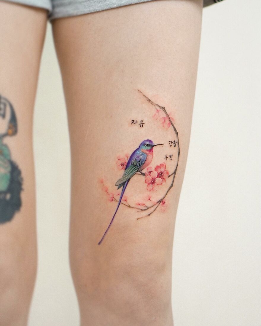 Tattoo of a colorful bird on a branch with blossoms, showcasing painting-like detail on a person's thigh.