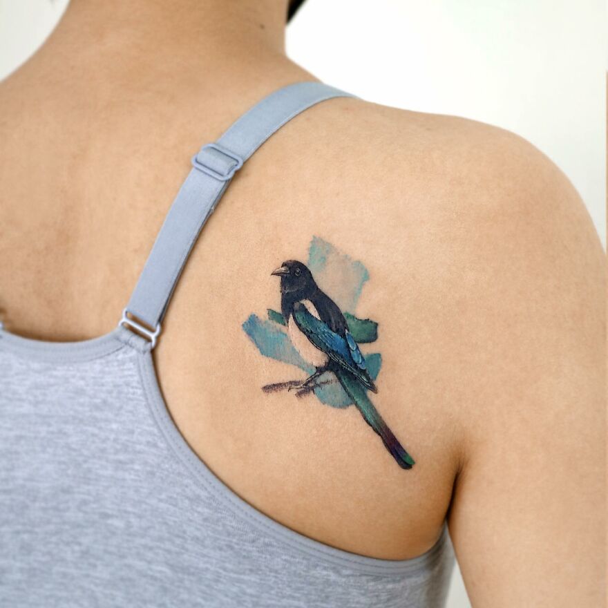Magpie tattoo on shoulder resembling a painting, featuring blue and black hues.