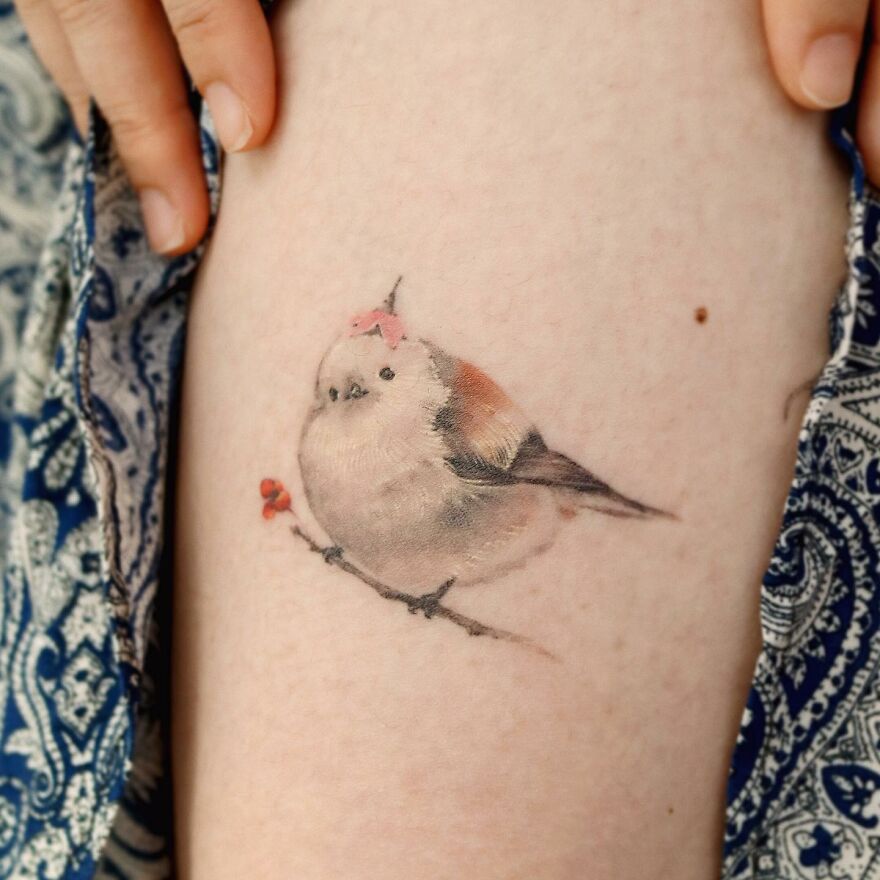 Painting-like tattoo of a small bird perched on a branch, resembling a delicate watercolor artwork on skin.