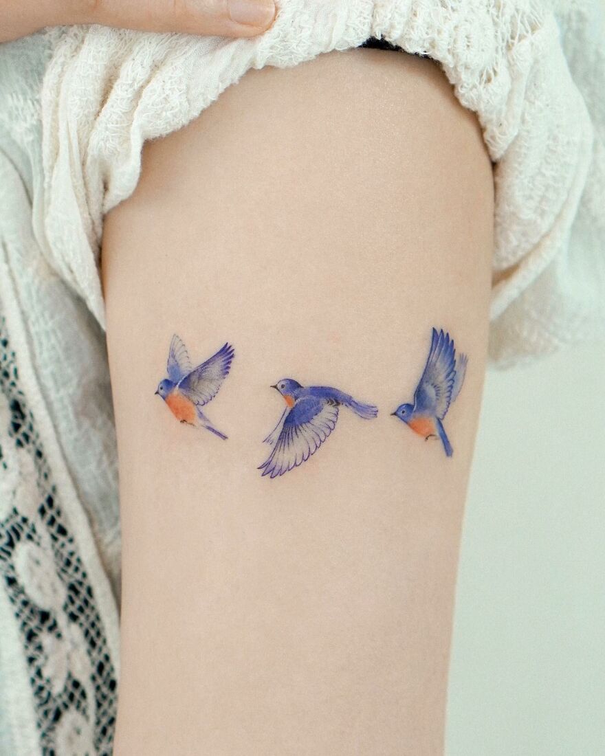 Painting-like tattoos of three vibrant blue and orange birds in flight on a person's skin.