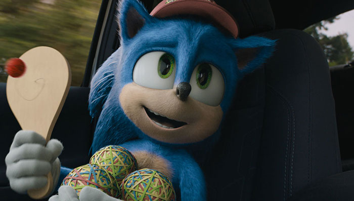 Cute animated character with blue fur and green eyes, holding a paddle and colorful balls, sitting in a car.