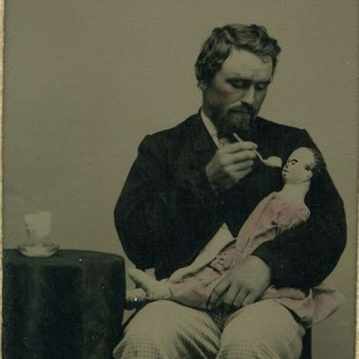 Hand-Coloured Photograph Of A Grown Man Feeding A Spooky Doll With A Spoon, C. 1864-66