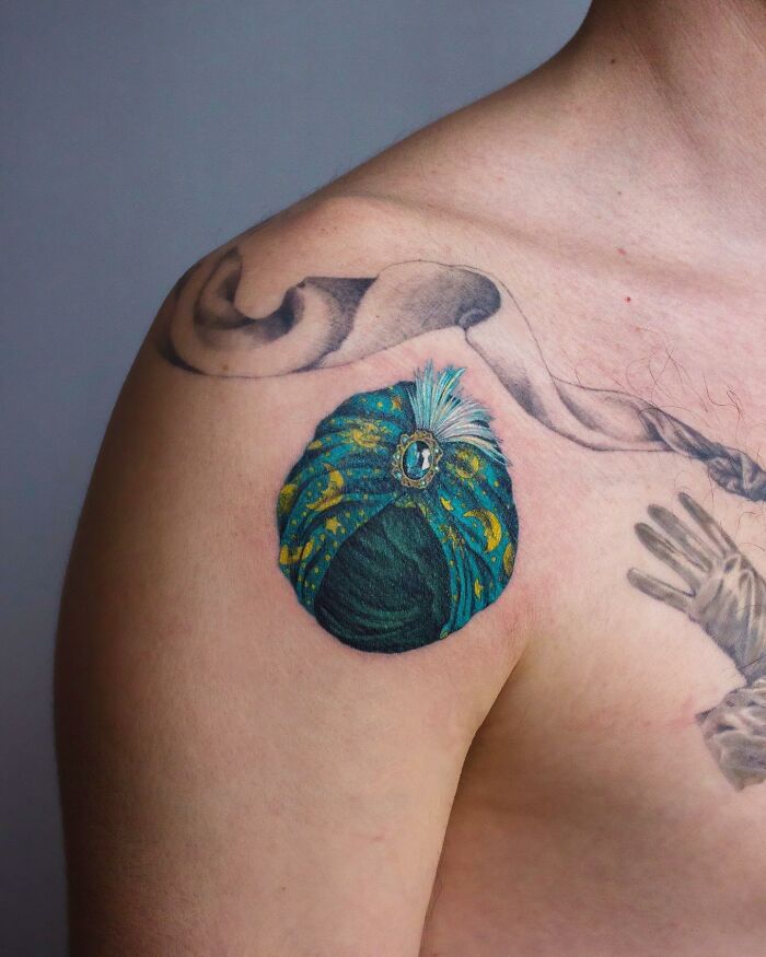 Colorful permanent jewelry tattoo on shoulder, featuring a detailed turban with a gemstone.