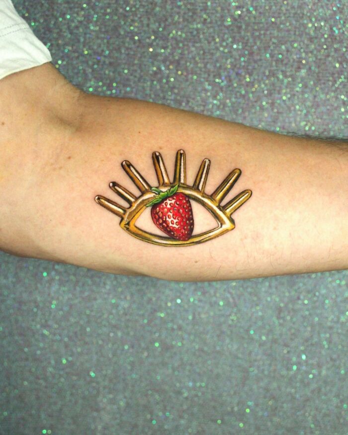 Tattoo of an eye with a strawberry instead of a pupil, showcasing permanent jewelry tattoo art on an arm.