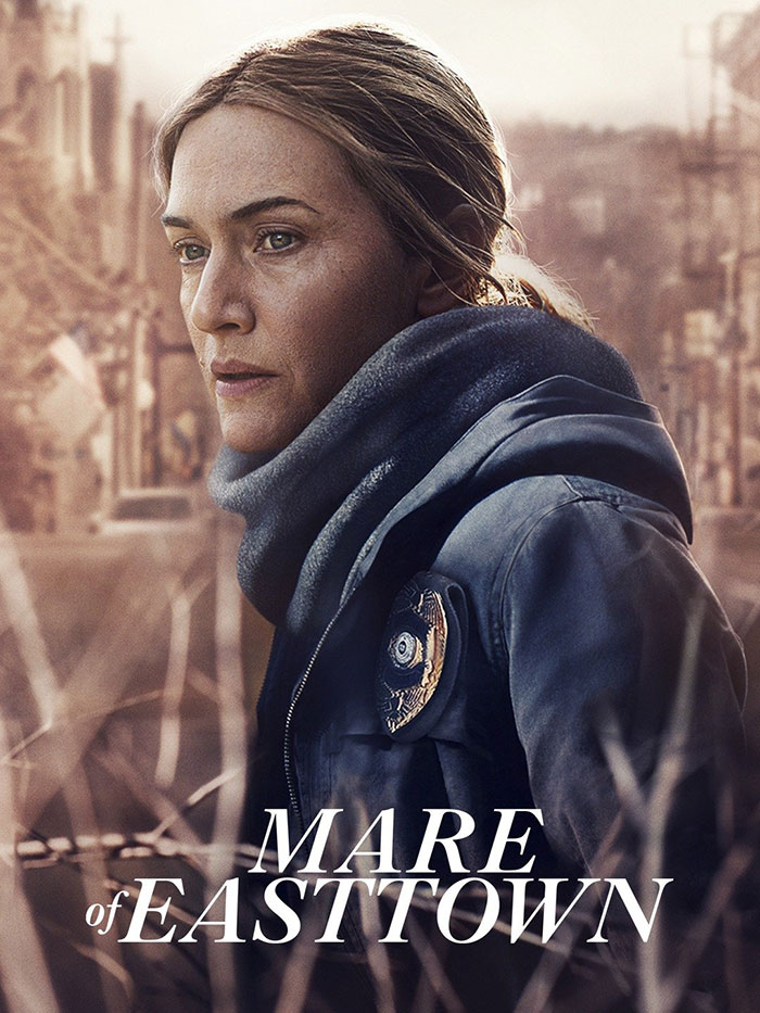 Woman in a dark coat and scarf, starring in popular HBO TV show "Mare of Easttown" with a blurred urban background.