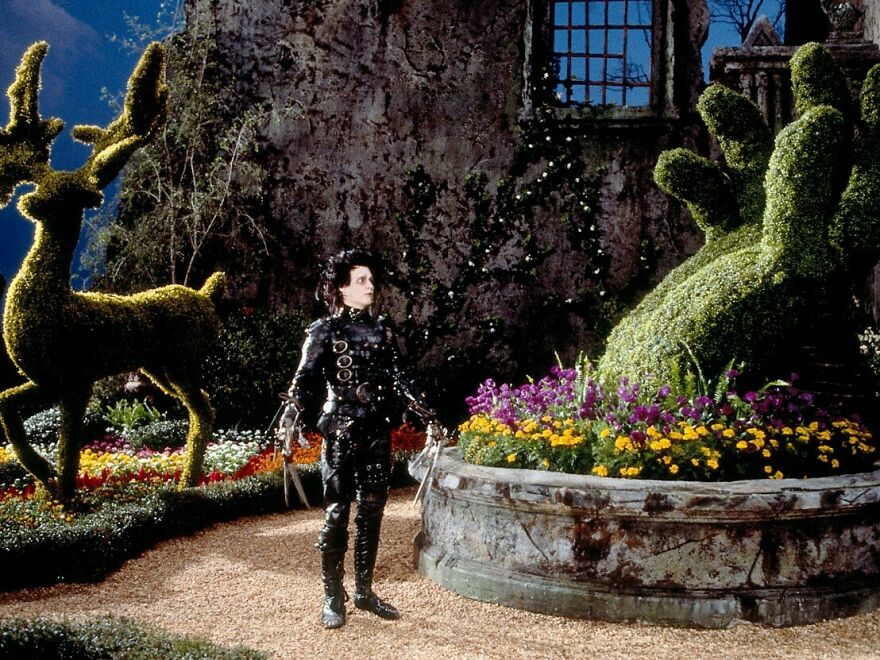Edward is outside in a park with many flowers and cut bushes like deer and a hand