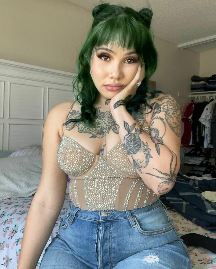 Woman with tattoos and green hair, sitting on bed, looking concerned. Woman with tattoos and green hair, sitting on bed, looking concerned.