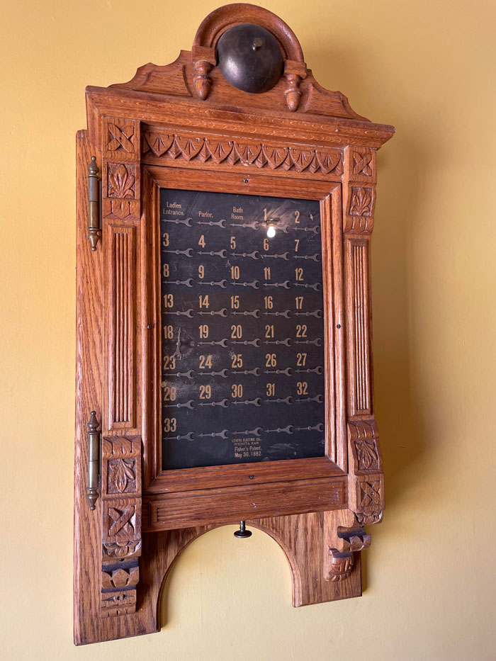 Wooden Machine Hanging On The Wall Of A Historical Building. It Features Dials That All Seem To Be In Sync. Knob Below When Pushed Makes The Dials Jump, But Turning Does Nothing. The Dials Are Numbered But A Few Are Labeled Parlor, Ladies Entrance And Bathroom