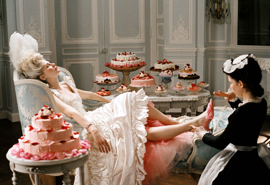 A woman in a cream dress relaxing in a chair surrounded by many cakes and a maid near her