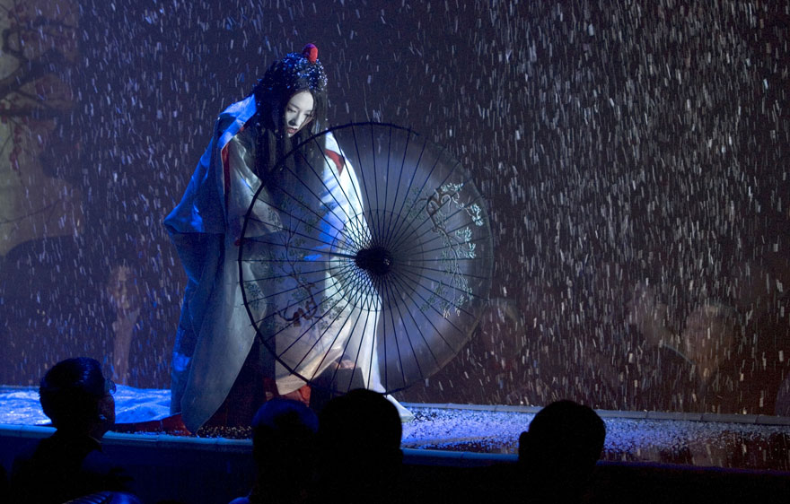 A Japanese girl in a kimono holding an umbrella stands on stage, and it's snowing 