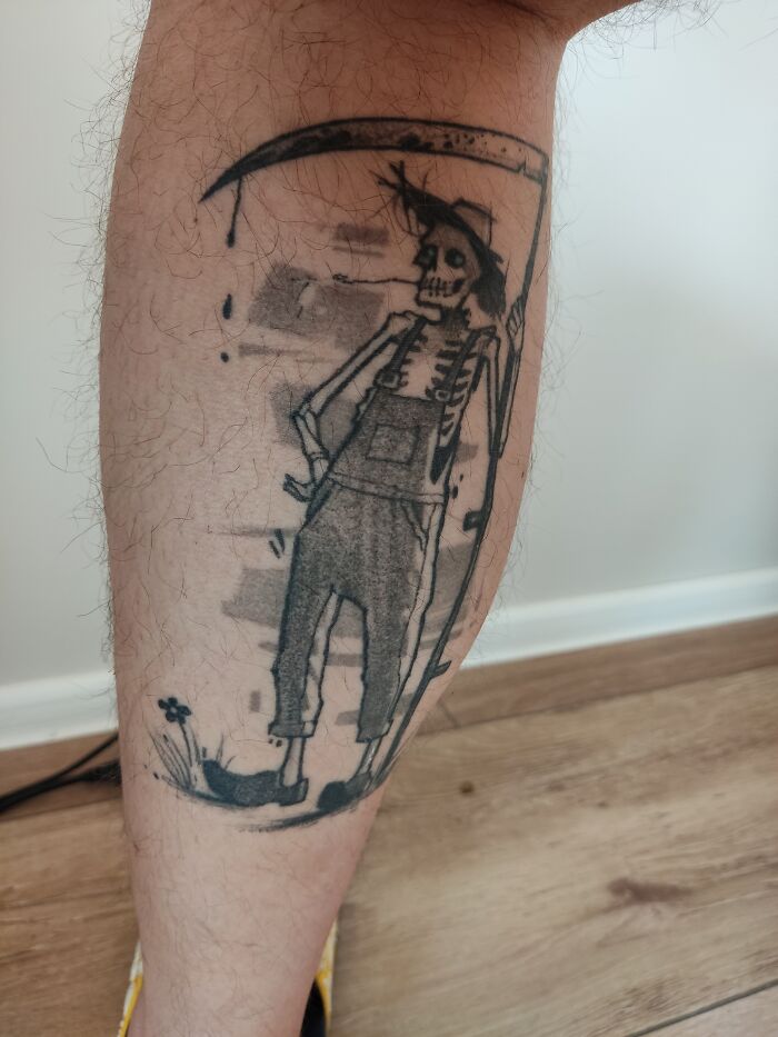 Tattoo of a skeleton wearing overalls and holding a scythe on a person's leg.