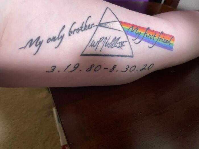 Tattoo on arm featuring a tribute with a triangle, rainbow, and dates, symbolizing a personal memorial.