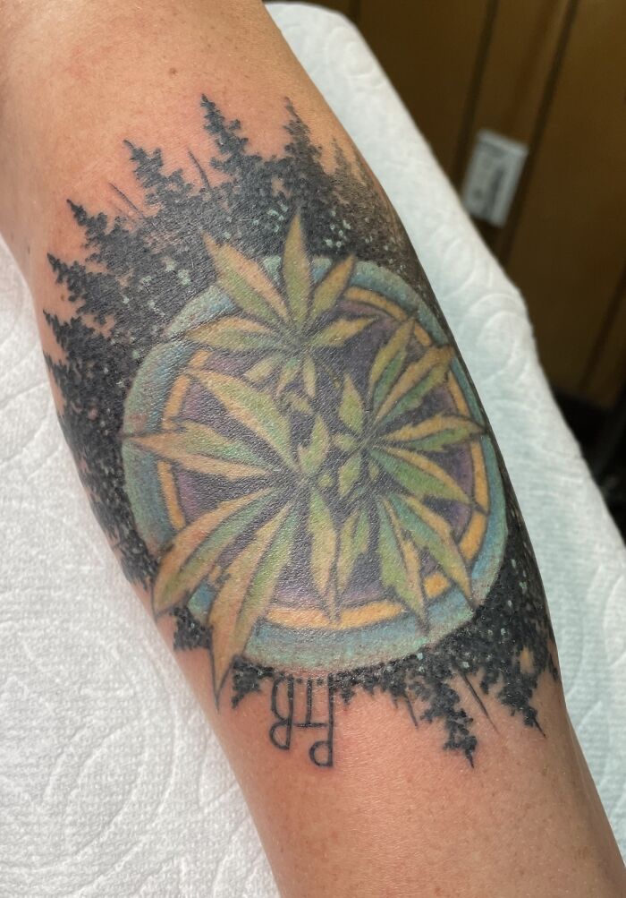 Tattoo with leaves and trees design on forearm, featuring intricate details in black and green, showcasing personal body art.