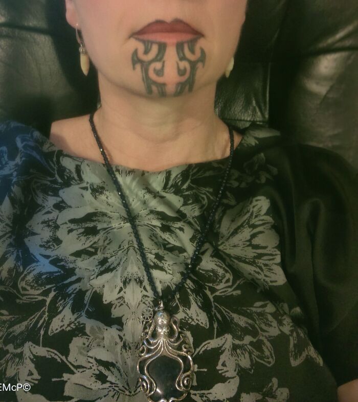 A person with a chin tattoo, wearing a floral top and a detailed necklace.