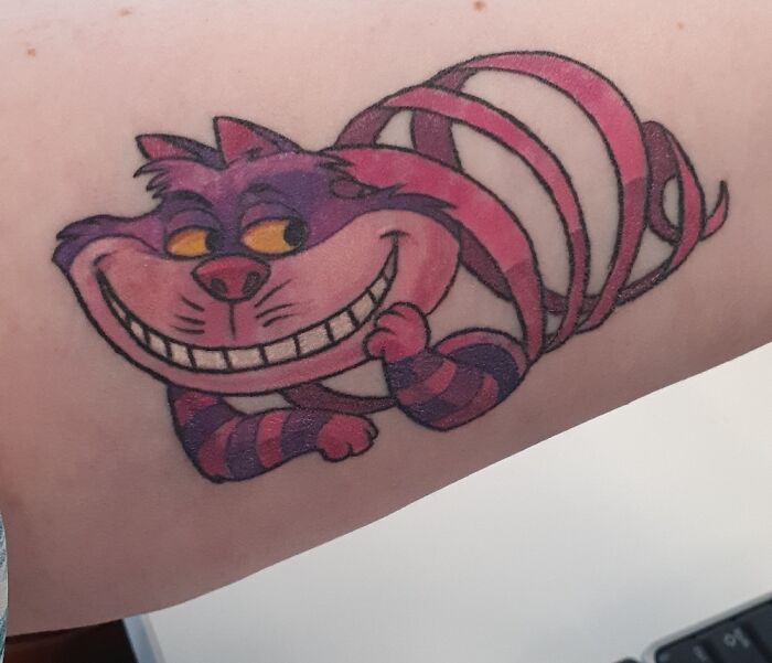 Colorful tattoo of a grinning purple and pink striped cat on an arm.