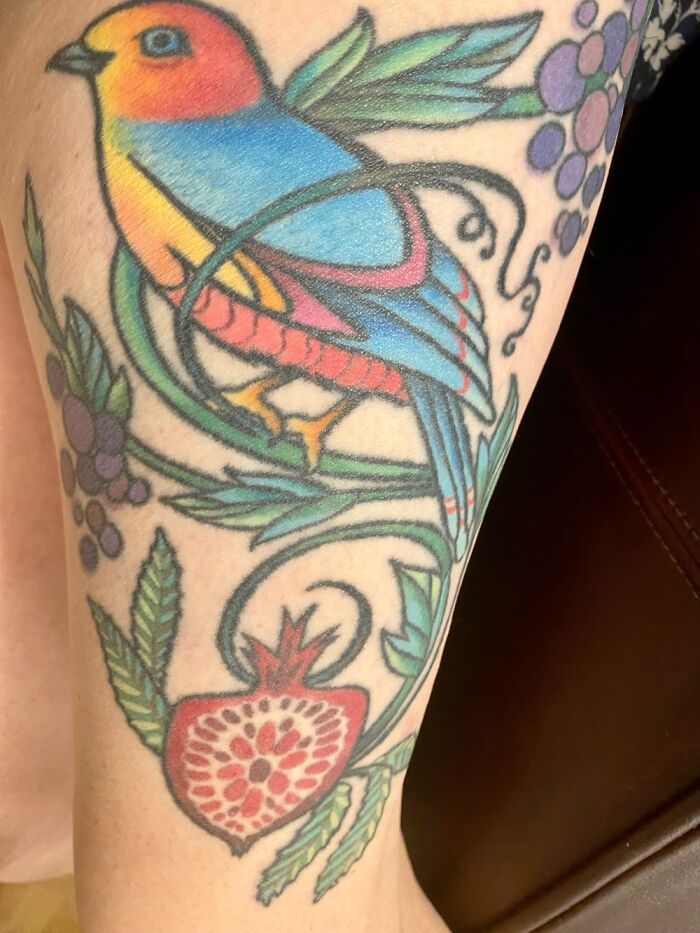 Colorful bird tattoo with floral and fruit designs, featuring a blue and orange bird on skin.