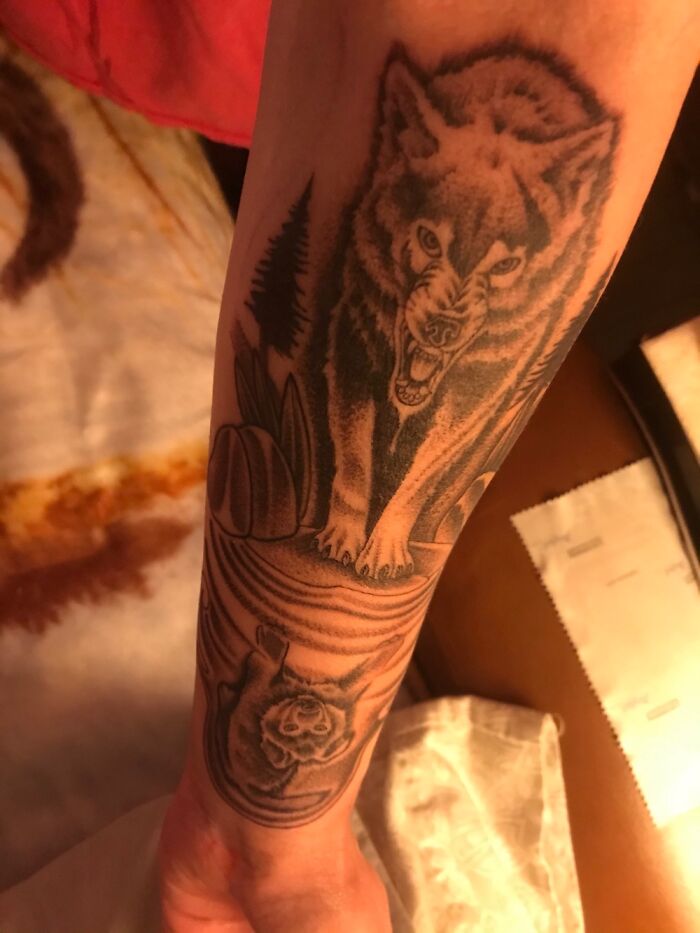 Wolf tattoo on forearm, detailed with forest background and reflection in water.