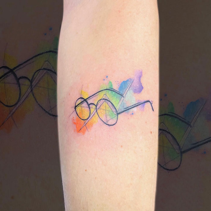 Minimalist Harry Potter tattoo with glasses and lightning bolt, watercolor style with rainbow hues on forearm.