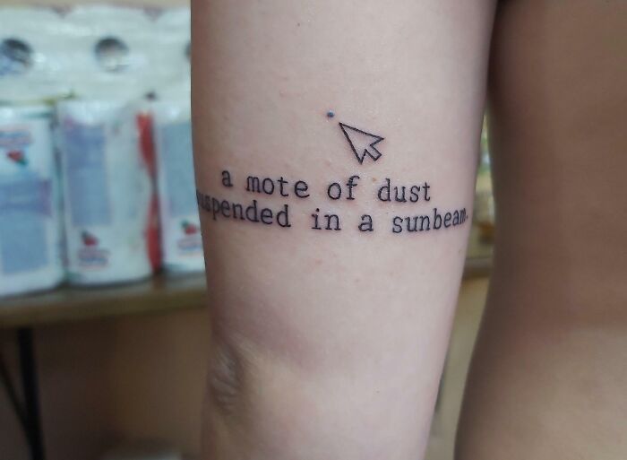 Word tattoo on arm reading "a mote of dust suspended in a sunbeam" with a small cursor icon.