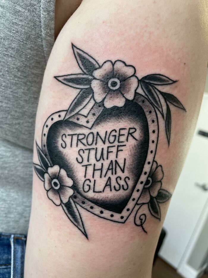 Tattoo of heart with flowers and text "Stronger Stuff Than Glass" on arm, highlighting meaningful word tattoos.