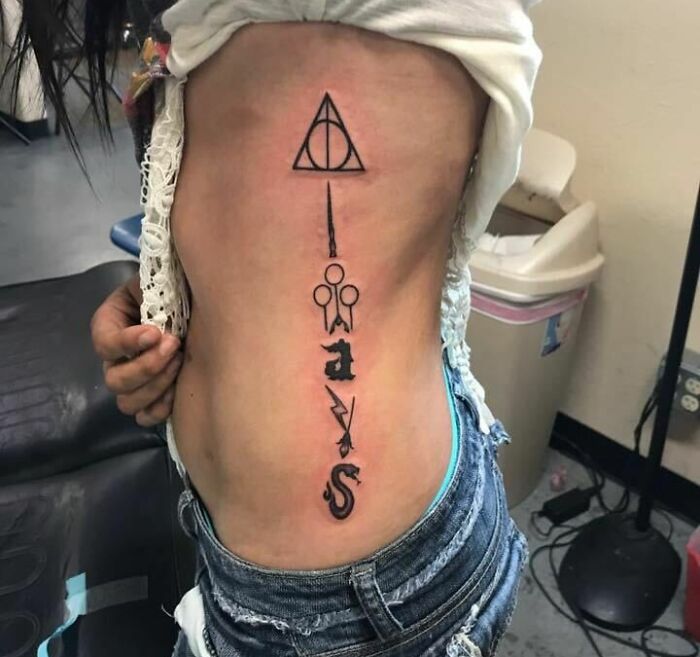 Harry Potter tattoo on a person's side featuring iconic symbols and text in black ink, wearing jeans and a white top.