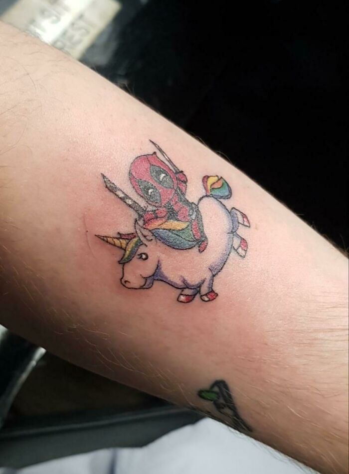 Marvel tattoo of a Deadpool superhero riding a unicorn on forearm.