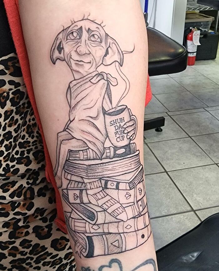 Harry Potter tattoo of Dobby sitting on books, holding a cup, on a person's arm.