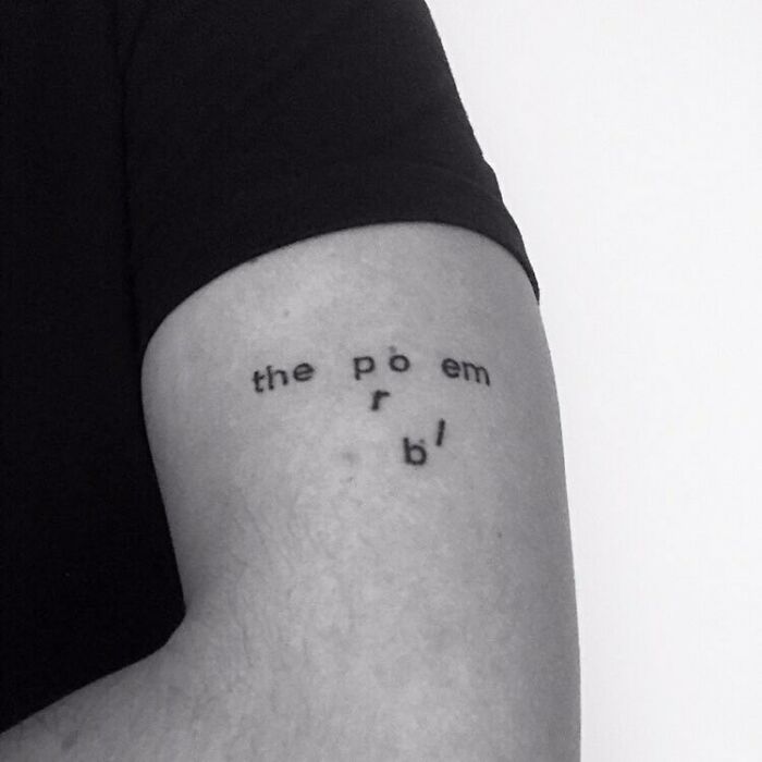 Minimalist word tattoo on arm in black ink, featuring scattered letters forming a phrase.