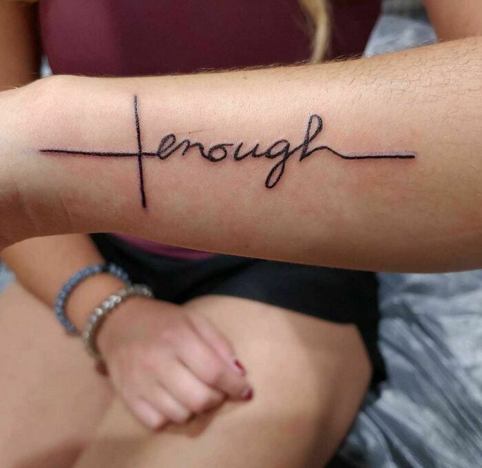 Tattoo of the word "enough" on a forearm, featuring a cross-like design.