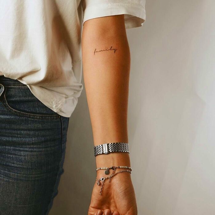 Minimalist word tattoo reading "family" on a person's forearm, paired with casual attire and a bracelet.