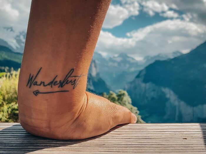 Word tattoo "Wanderlust" on ankle with mountain view in background.